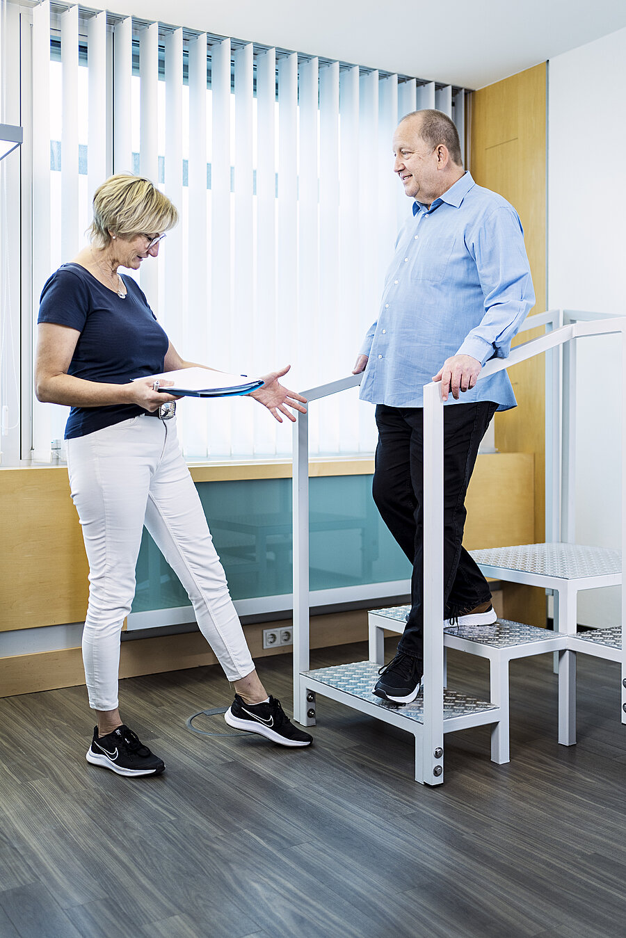 Ein Mann steigt eine kleine Treppe in einem Büro herab. Eine Frau in weißer Hose steht vor ihm. Sie hat ein Heft in der Hand und zeigt auf seine Füße.